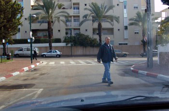 גבר חוצה במעבר חצייה מסומן ברחוב מגורים, בין רכבים חונים.